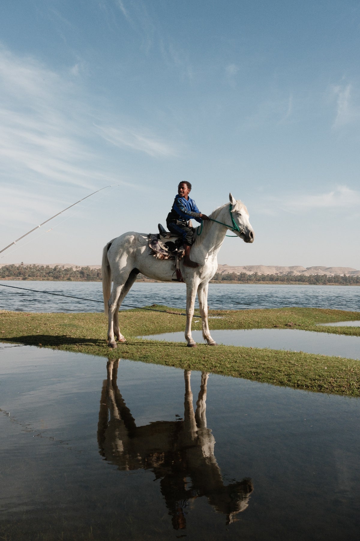 Hamza riding his white horse on a strip of grass by the Nile, the horse reflected in the water below