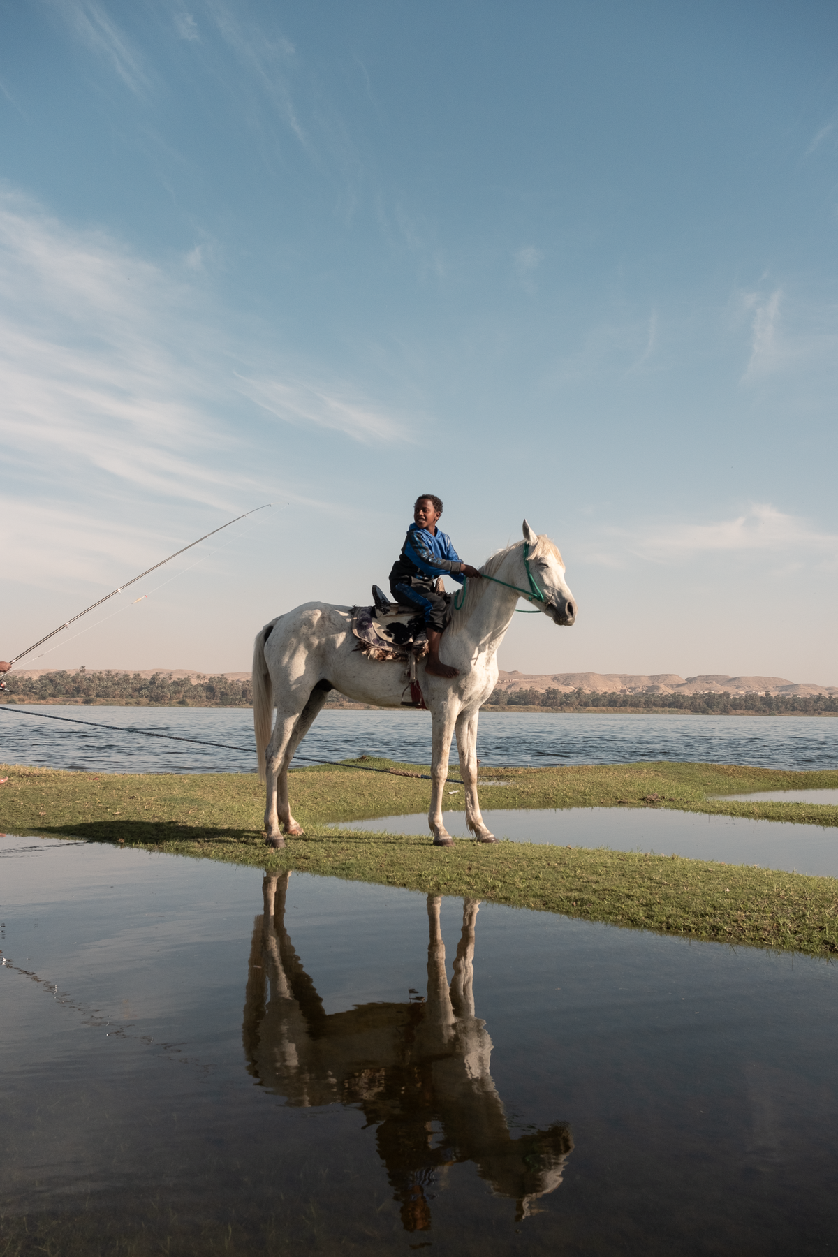 Hamza riding his white horse on a strip of grass by the Nile, the horse reflected in the water below