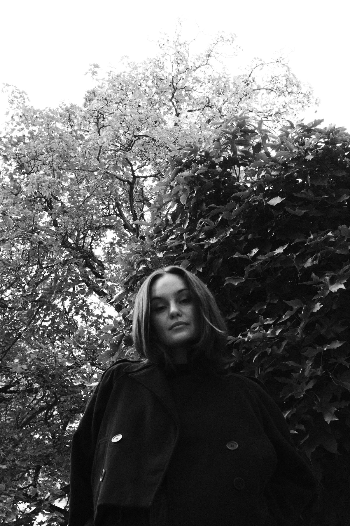 Helena Wedekind from below in a dark coat, looking down at the camera, trees behind her