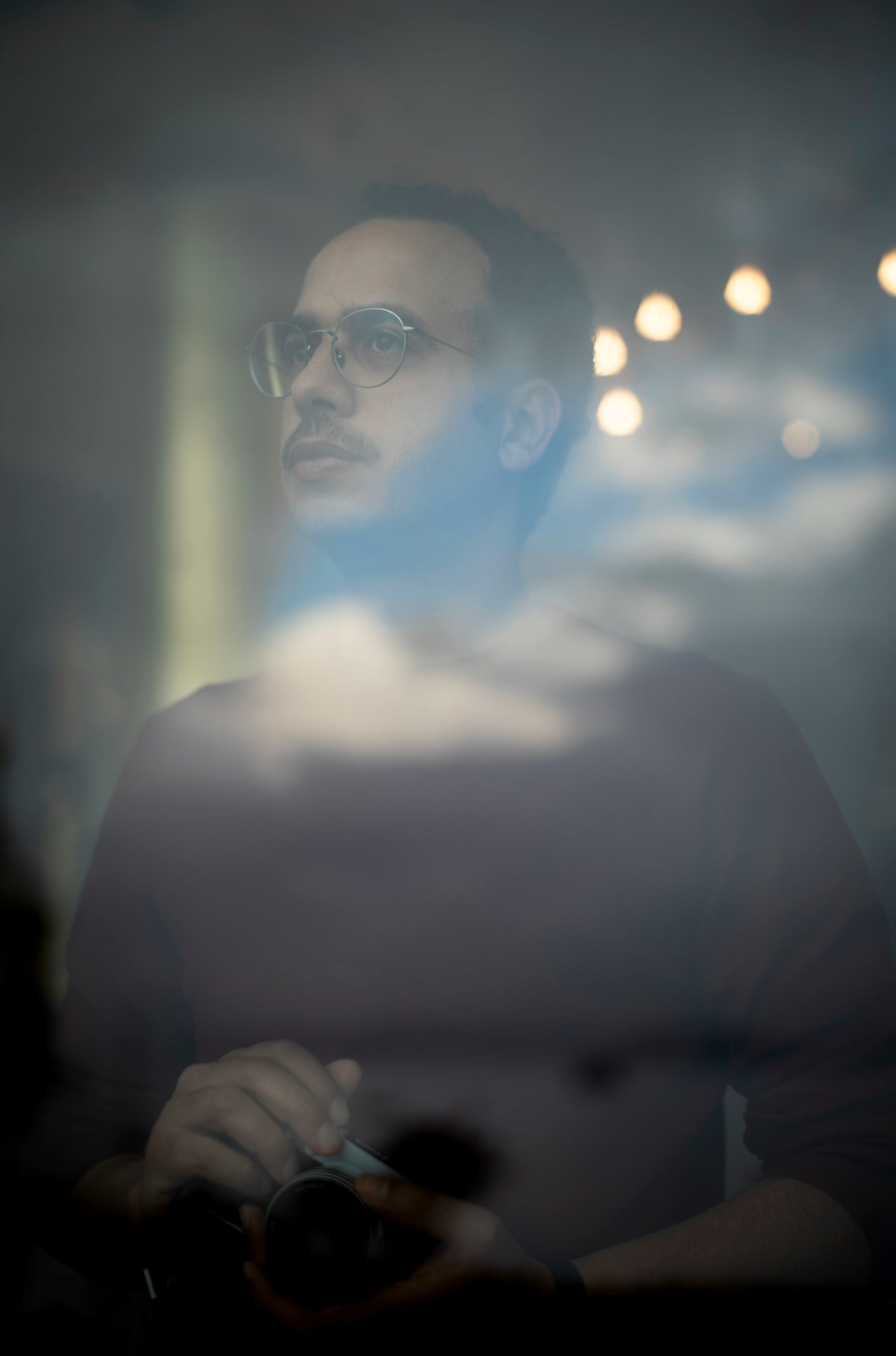 Portrait of photographer Ahmed Kamal Ali holding a camera, seen through glass with cloud reflections