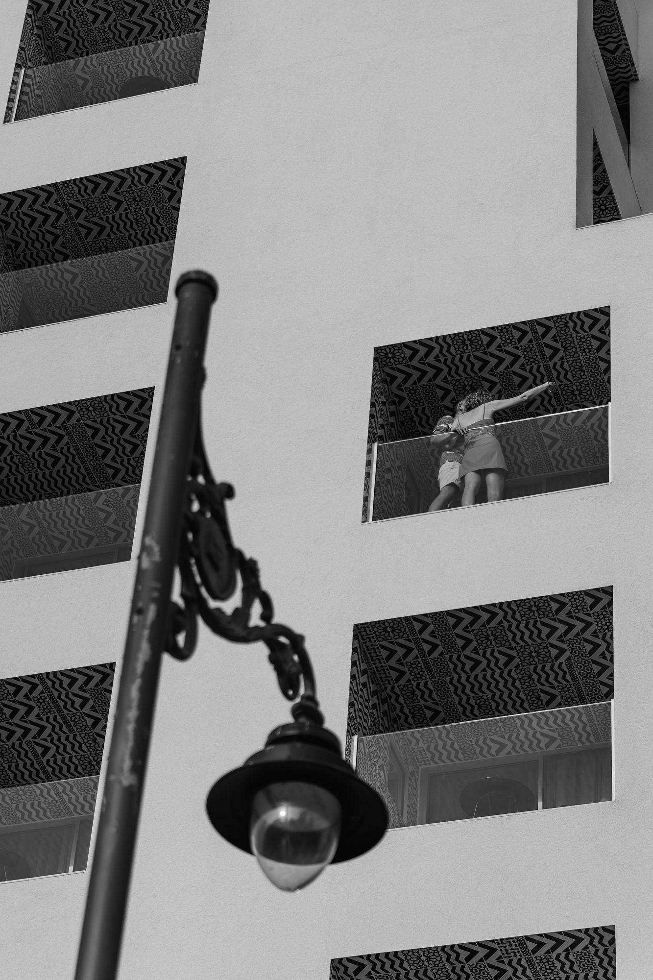Black and white street view in Málaga, a street lamp in the foreground and a person on a hotel balcony